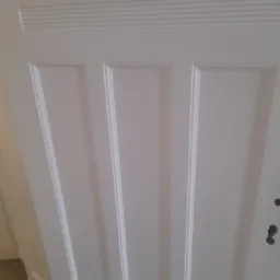 Białe, pomalowane drzwi wewnętrzne z charakterystycznymi frezowaniami i panelami, widoczne ślady po zamku i klamce, w tle fragment podłogi zabezpieczony folią malarską.