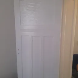 Białe, klasyczne drzwi wewnętrzne z górnym panelem ze szkła ornamentowego, świeżo pomalowane, widoczne zawiasy i fragment ościeżnicy