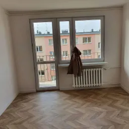 Pokój po remoncie z parkietem ułożonym we wzór jodełki, widok na balkon i budynek za oknem, na parapecie wisi kurtka.