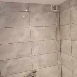 Ściana łazienki wyłożona szarymi, matowymi płytkami w dużym formacie, z widoczną kratką wentylacyjną w rogu i baterią prysznicową w dolnej części kadru.