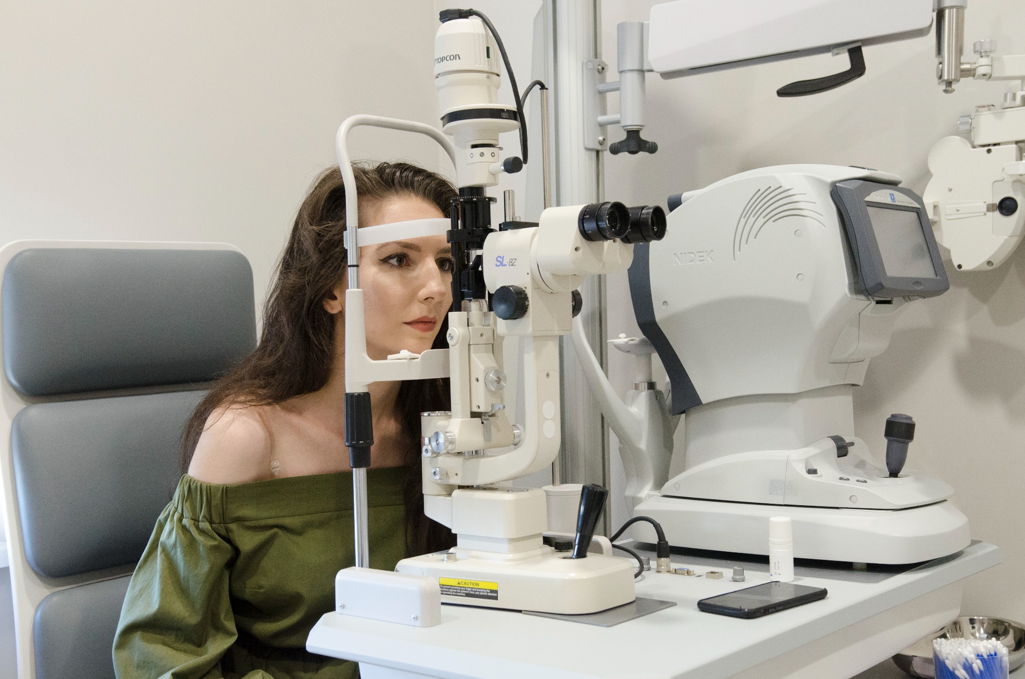 Kobieta z brązowymi włosami poddawana badaniu wzroku przy użyciu lampy szczelinowej Nidek SL-8Z, w tle widoczny aparat do pomiaru parametrów rogówki.