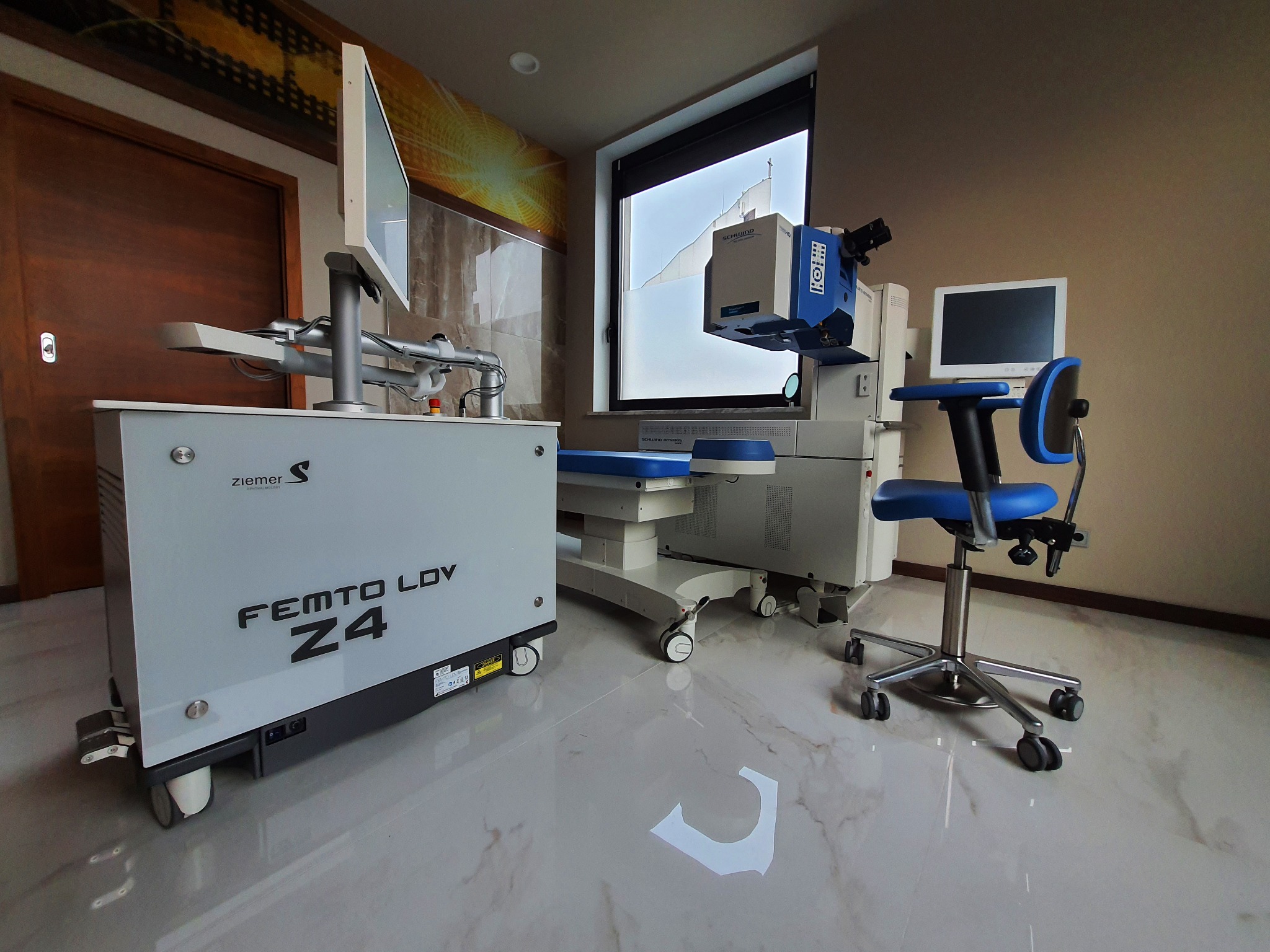Nowoczesny gabinet z zaawansowanym sprzętem okulistycznym: laser femtosekundowy Ziemer Femto LDV Z4 i laser Schwind Amaris, fotel zabiegowy na kółkach, monitor i marmurowe płytki na podłodze.