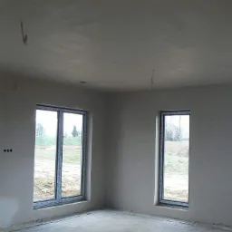 Wnętrze surowego domu z trzema oknami po zakończeniu prac na ścianach i suficie, widoczne wystające kable elektryczne.