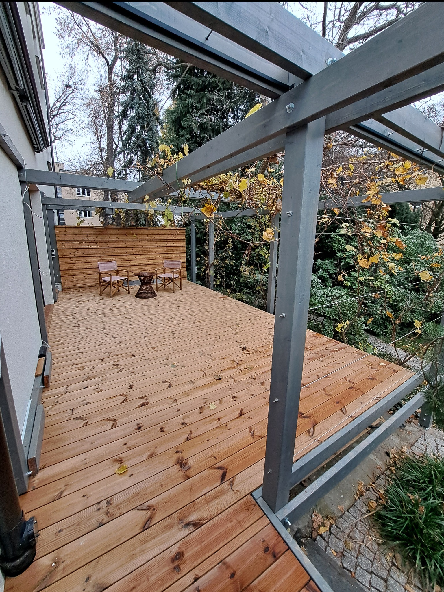 Drewniany taras z szarą pergolą i drewnianą ścianką. Dwa krzesła i stolik na tle zieleni. Jesienne liście na pergoli. Widok na budynek w tle.