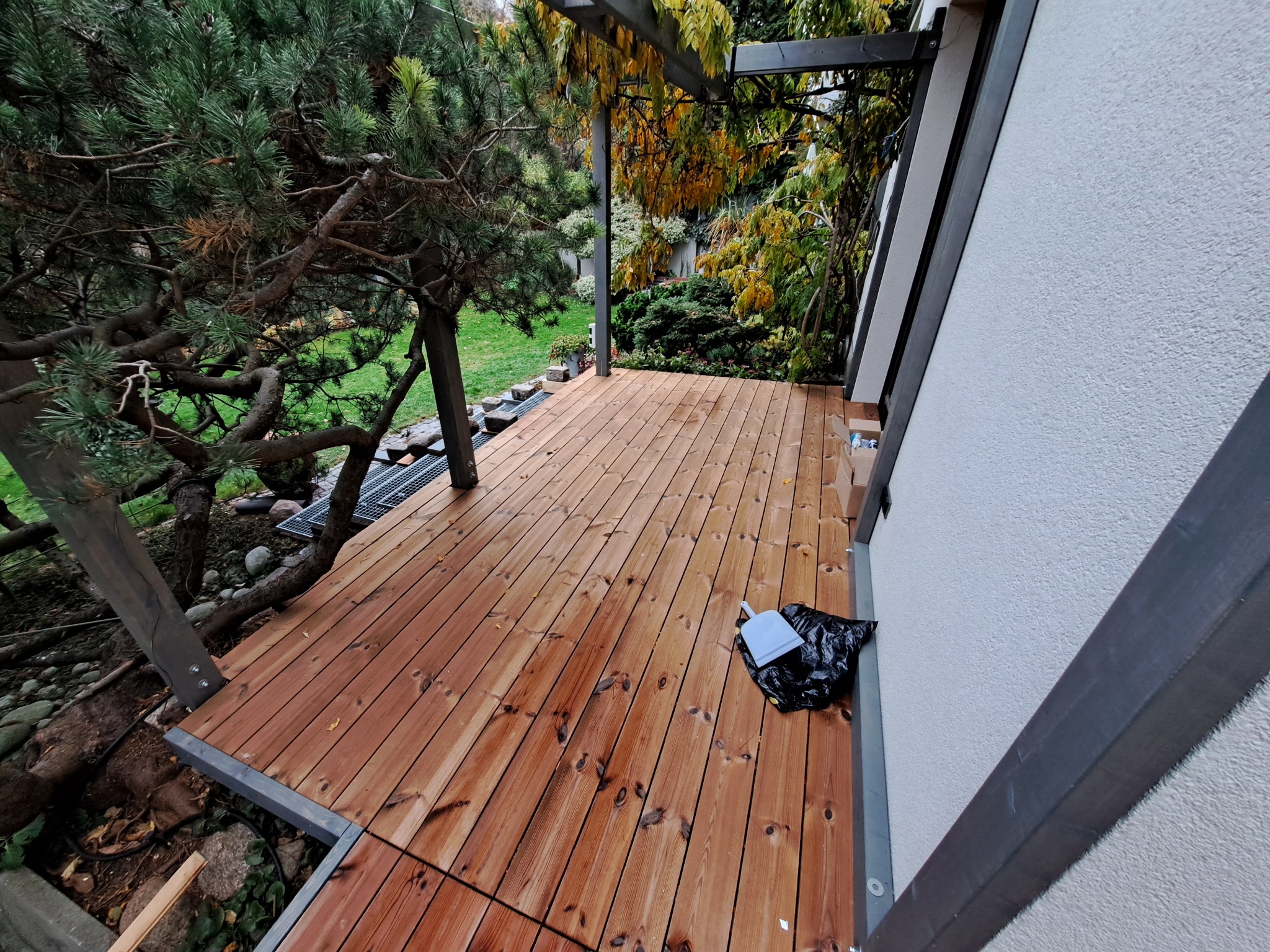 Nowy taras z desek w odcieniach brązu, z elementami konstrukcyjnymi w kolorze szarym, otoczony zielenią ogrodu. Widoczny fragment ściany budynku.