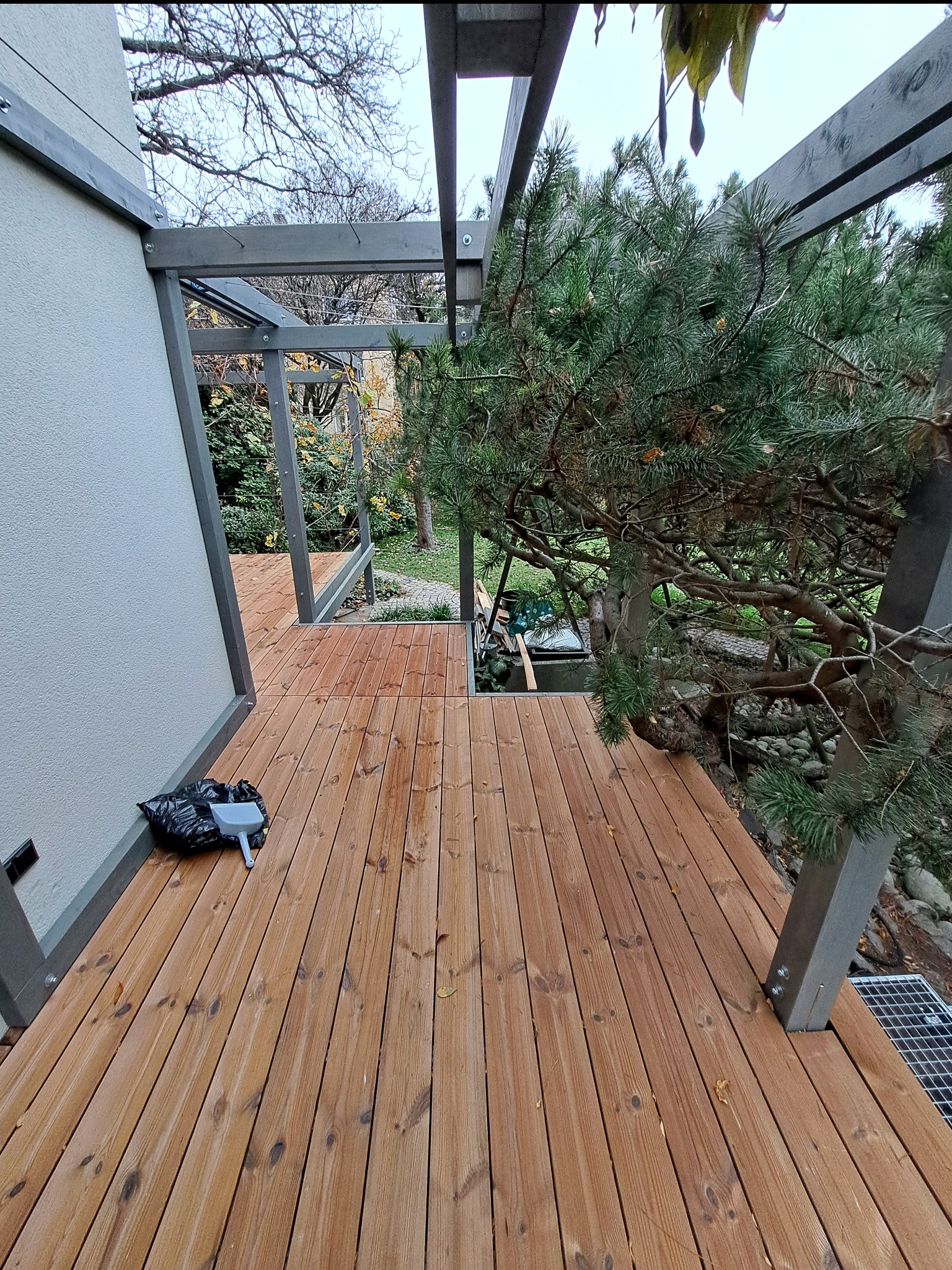 Nowoczesny taras drewniany z metalową konstrukcją, widok z góry, z elementami zieleni i workiem na śmieci w rogu. Deski ułożone równolegle, widoczne mocowania.