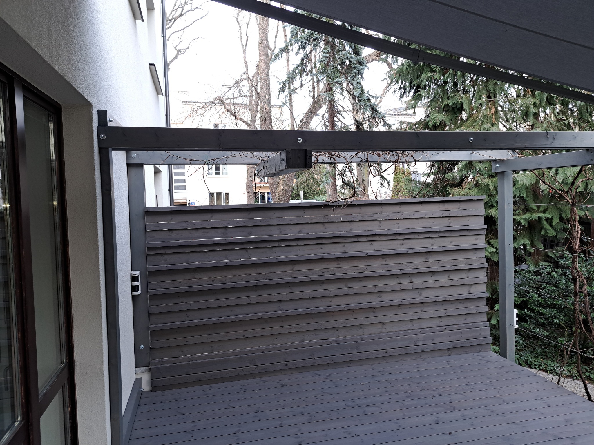 Nowoczesny, szary taras z drewnianą podłogą i ażurową ścianką, zadaszony pergolą. Widok na zieleń ogrodu. Minimalistyczny design.