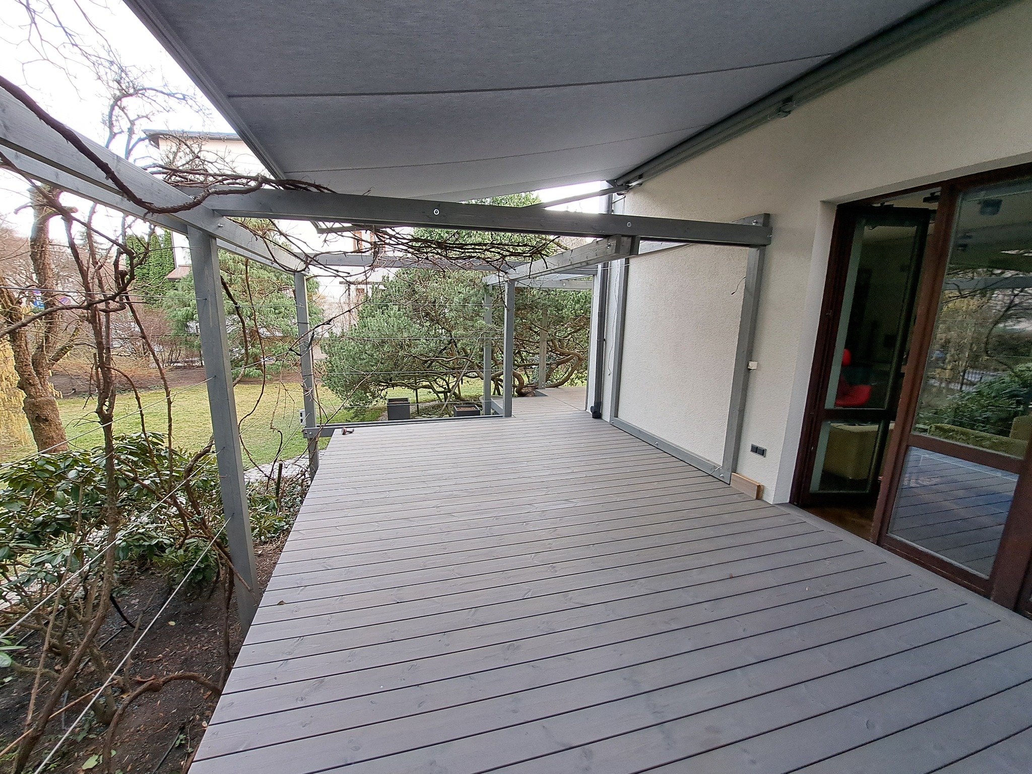 Zadaszony taras z szarych desek, metalowa konstrukcja z pnączami, widok na ogród. Nowoczesny design, zintegrowany z naturą, drzwi balkonowe z ciemnego drewna.