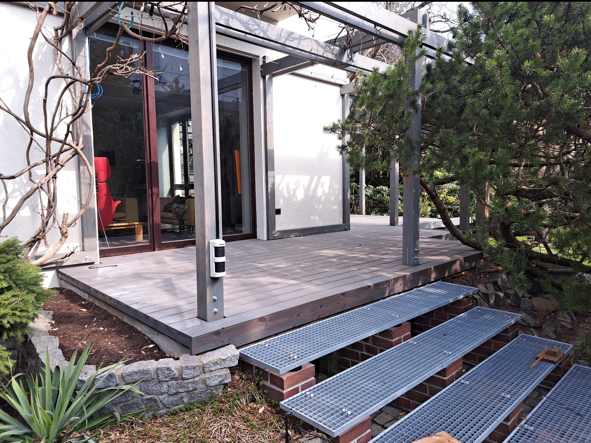 Nowoczesny taras z szarego drewna z metalowymi schodami kratowymi, otoczony zielenią i elementami architektury ogrodowej. Widoczne detale konstrukcyjne i roślinność.