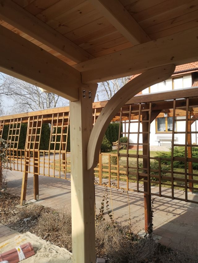 Drewniana konstrukcja z kratownicą w ogrodzie, widok z dołu, jasne drewno, elementy złączne, częściowo zadaszona, w tle dom z białymi oknami.