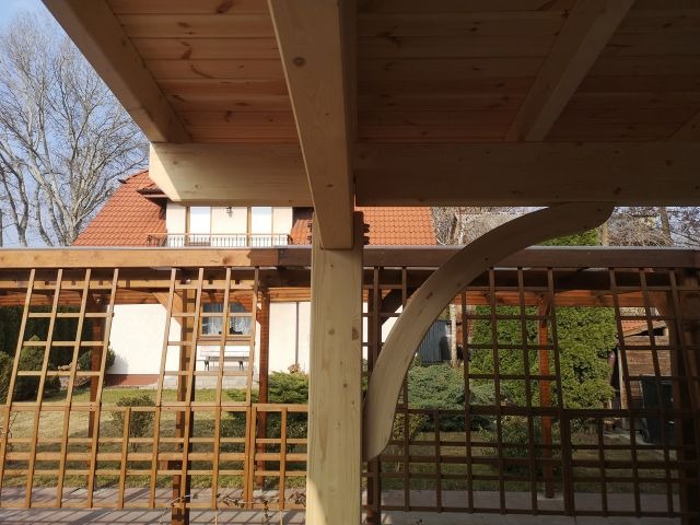 Drewniana konstrukcja tarasu z kratownicą, widok z wnętrza. Jasne drewno, łukowate wsporniki, w tle dom z czerwonym dachem i zielony ogród. Słoneczny dzień.