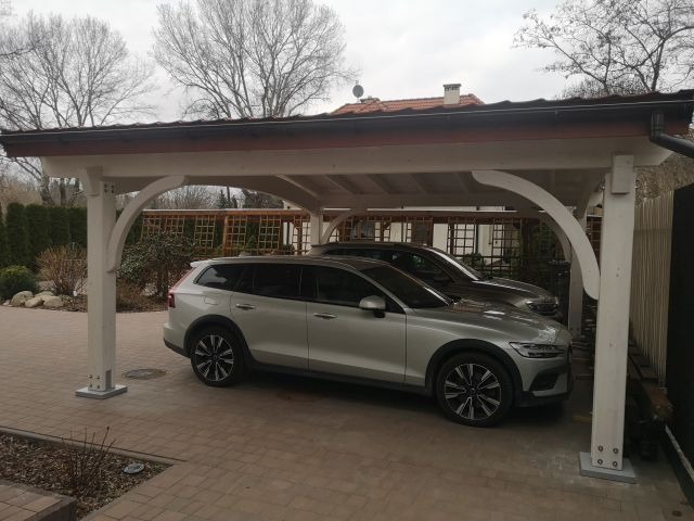 Zadaszone miejsce parkingowe z białymi słupami i drewnianą konstrukcją dachu, pod którym stoją dwa samochody. Kostka brukowa na podłożu.