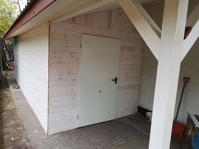 Biała, drewniana konstrukcja z drzwiami i wspornikami, widoczna z boku. Czerwona łopata oparta o ścianę. Widoczne ślady użytkowania i niedoskonałości w malowaniu.