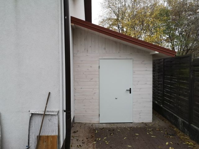 Biały, drewniany domek narzędziowy z brązowym dachem i białą, gładką bramą, stojący obok białego budynku i ciemnego ogrodzenia na brukowanym podłożu.