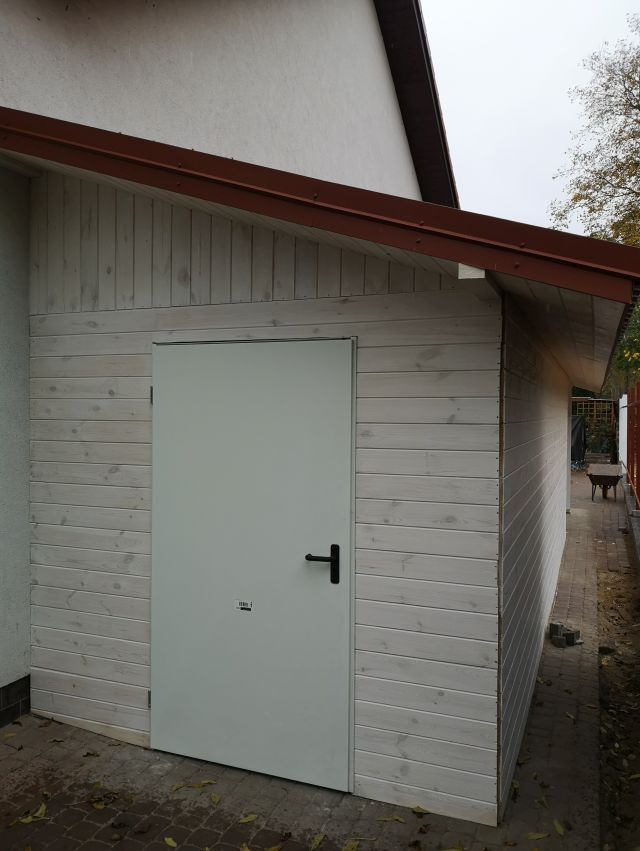 Drewniany budynek gospodarczy z białymi deskami i brązowym dachem, szara, metalowa brama z czarną klamką. Widok z zewnątrz, jesienne liście na ziemi.