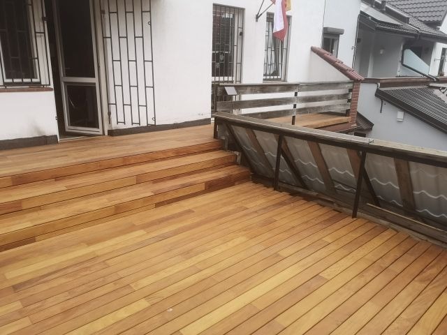 Jasny, drewniany taras z balustradą, prowadzący do budynku z oknami z kratami. Widoczny fragment dachu sąsiedniego budynku i flaga na budynku.