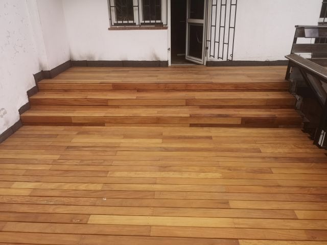 Jasnobrązowy taras z drewna egzotycznego, prowadzący schodami do wejścia. Widoczne detale desek i metalowa barierka. Minimalistyczny styl, naturalne światło.