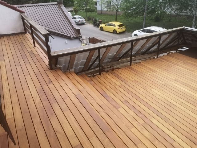 Wykończony taras z desek z drewnianą balustradą, widok z góry, w tle zabudowa miejska i zaparkowane samochody. Deski ułożone równolegle, ciepły odcień drewna.