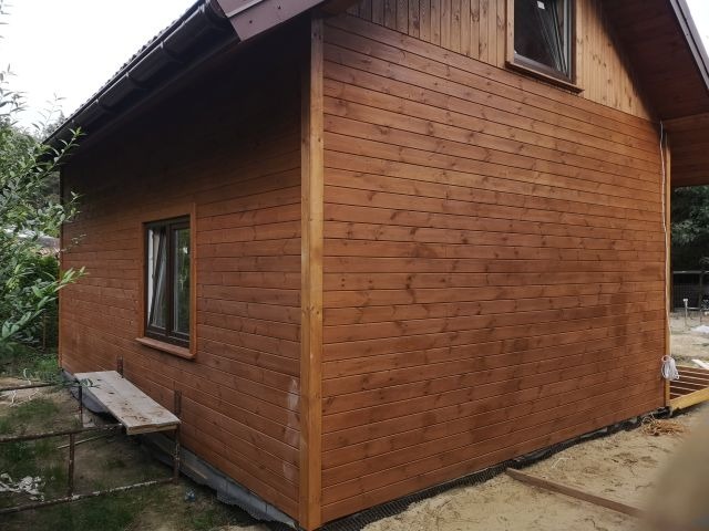 Drewniany domek letniskowy z brązową elewacją z desek, widoczny z zewnątrz, z małym oknem i fragmentem dachu. W tle zieleń drzew.