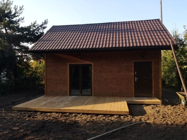 Drewniany domek z brązowym dachem i jasnym tarasem. Widoczne drzwi i okno. Otoczenie w trakcie prac ziemnych, z widoczną taczką i wężem ogrodowym.