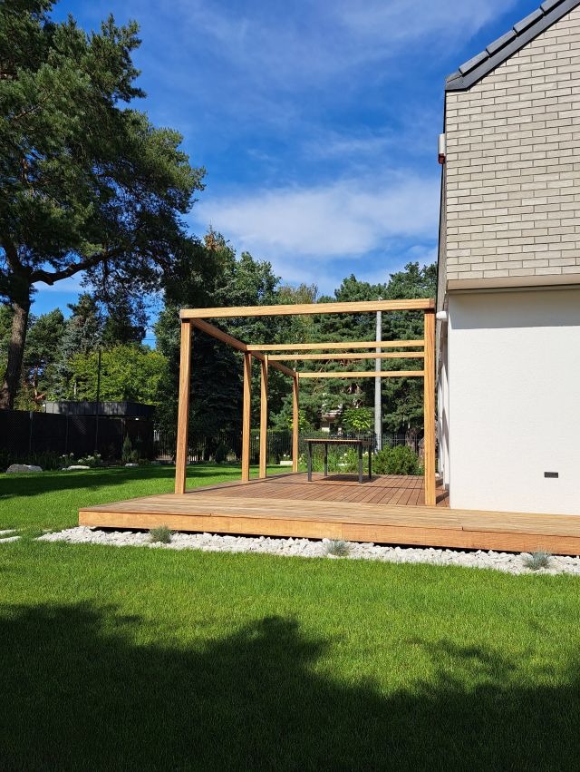 Drewniany taras z pergolą w ogrodzie, Warszawa. Jasne deski, białe kamienie dekoracyjne, zielony trawnik i błękitne niebo tworzą harmonijną przestrzeń.