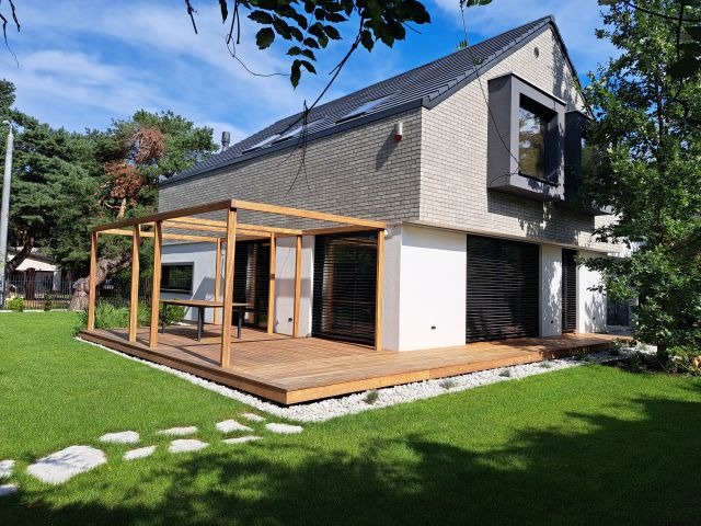 Nowoczesny dom z drewnianym tarasem i pergolą, otoczony zielonym trawnikiem i ścieżką z kamieni. Jasne drewno tarasu kontrastuje z bielą ścian budynku.