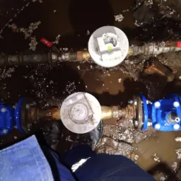 Dwa r&oacute;wnoległe rurociągi z zaworami i licznikami, zanurzone w błocie, widoczne z g&oacute;ry, z fragmentem niebieskiego kombinezonu roboczego w dolnej części kadru.