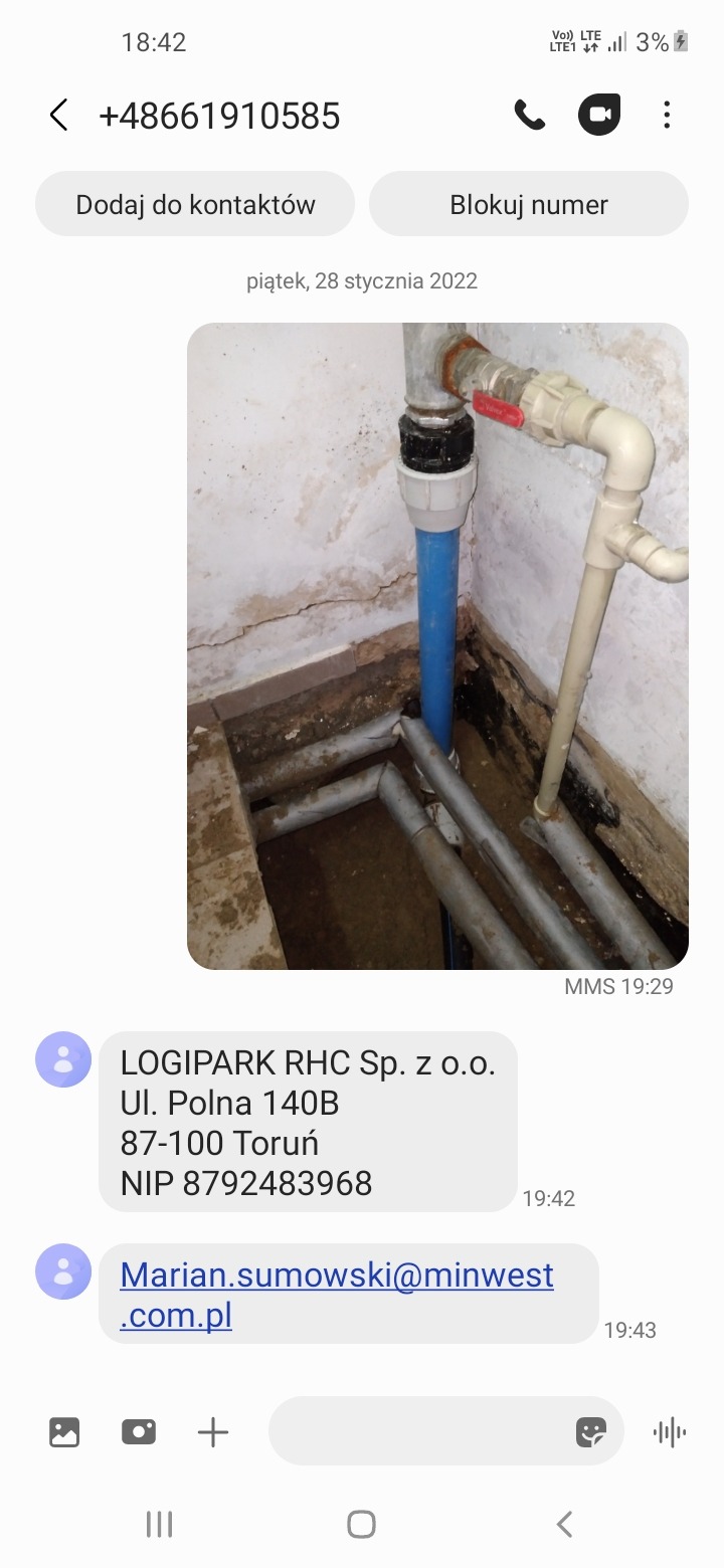 Zdjęcie z telefonu przedstawiające rury wodociągowe w piwnicy, widoczne połączenia z zaworem, ściany z zaciekami, rury częściowo w ziemi, zdjęcie niskiej jakości przesłane jako MMS.