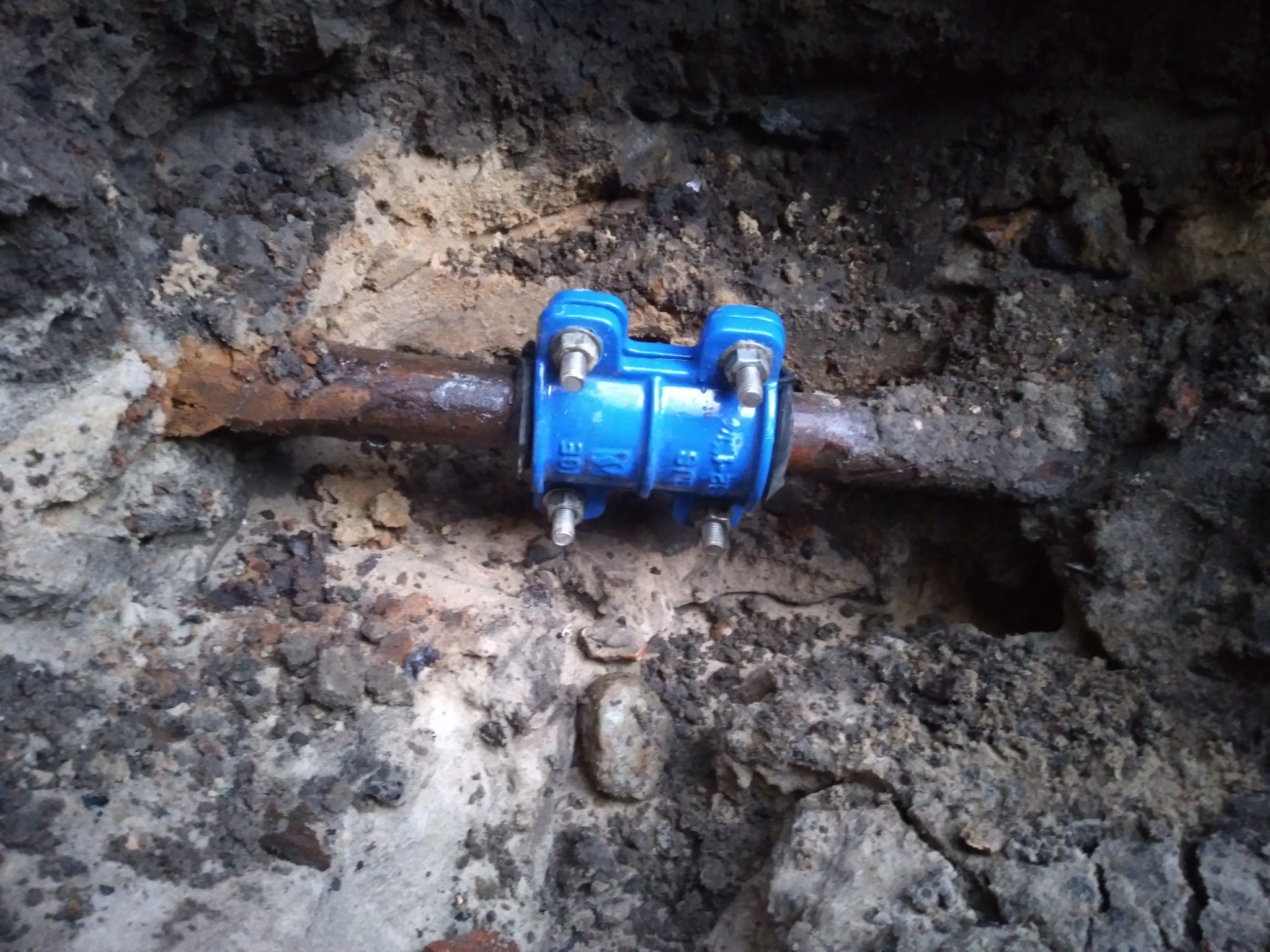 Zardzewiała rura wodna w wykopie, zabezpieczona niebieską obejmą z nakrętkami, otoczona ziemią i gruzem.