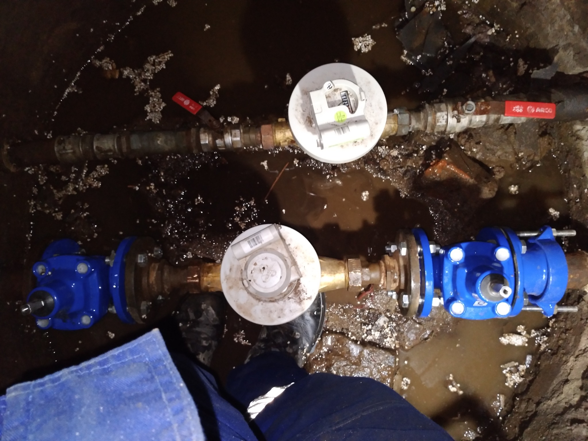 Dwa równoległe rurociągi z zaworami i licznikami, zanurzone w błocie, widoczne z góry, z fragmentem niebieskiego kombinezonu roboczego w dolnej części kadru.