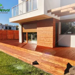 Nowoczesny dom z tarasem drewnianym na kilku poziomach, z drewnianą ścianą i pergolą, widok z trawnika w słoneczny dzień.