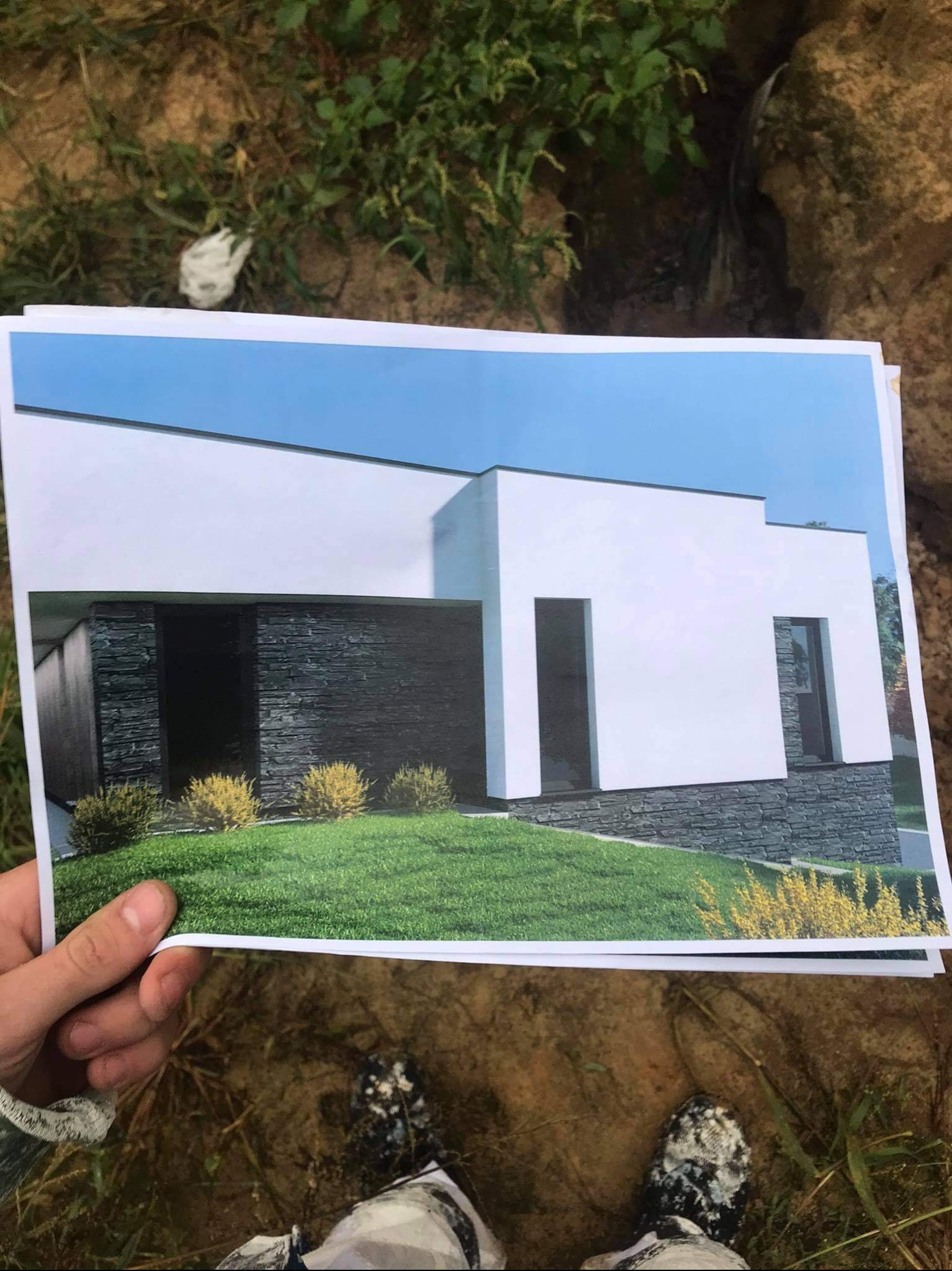 Osoba trzyma w dłoni wydrukowane zdjęcie nowoczesnego domu z białymi ścianami i elementami z ciemnego kamienia, na tle ziemi i zieleni.