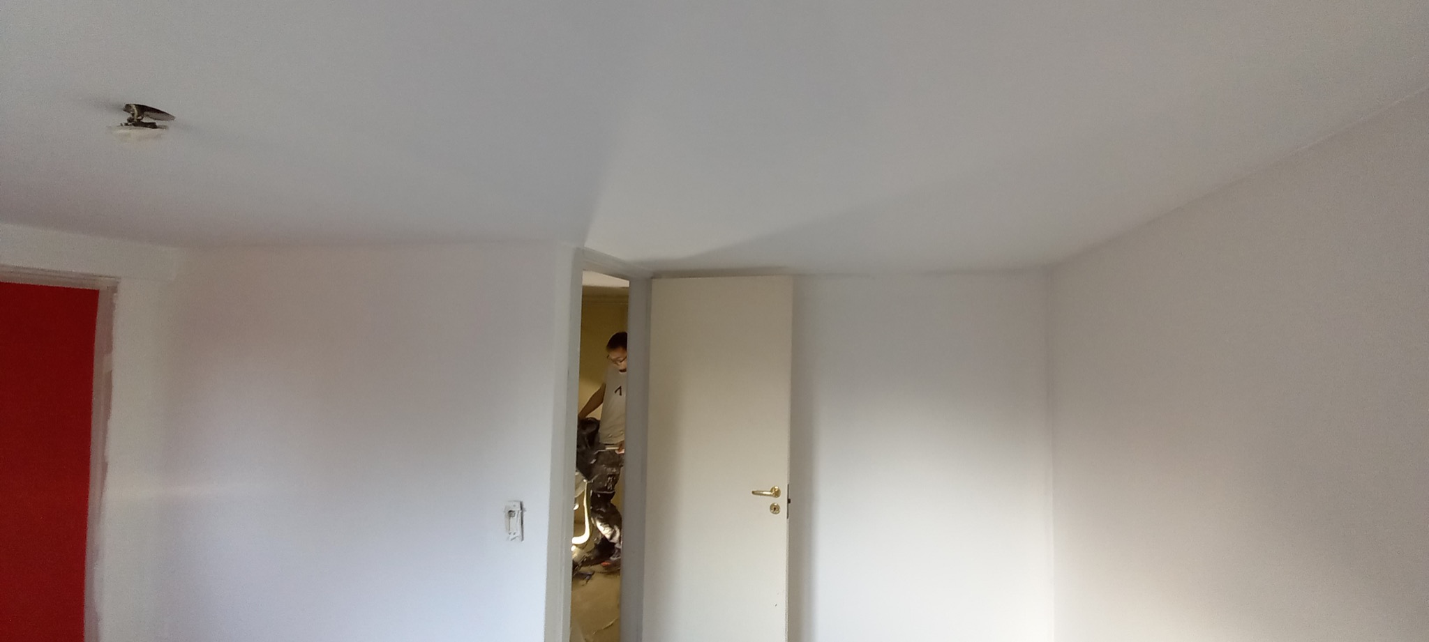 Świeżo pomalowane na biało wnętrze pokoju z widocznym fragmentem czerwonej ramy drzwi po lewej stronie i białymi drzwiami po prawej, przez otwarte drzwi widać malarza podczas pracy.