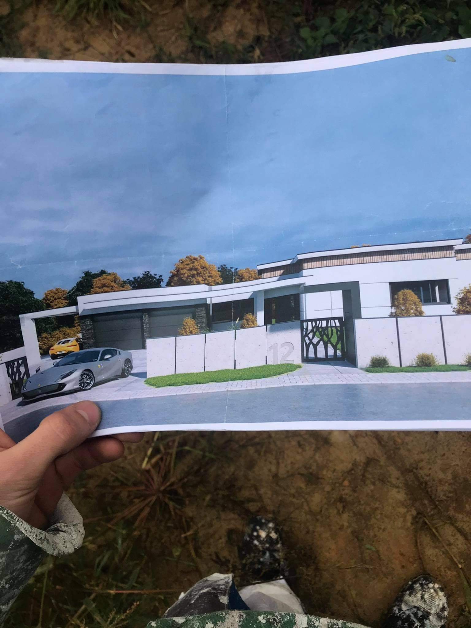 Ręka trzyma wydrukowany projekt nowoczesnego domu z garażem na dwa samochody i ozdobną bramą, widoczny srebrny samochód sportowy.