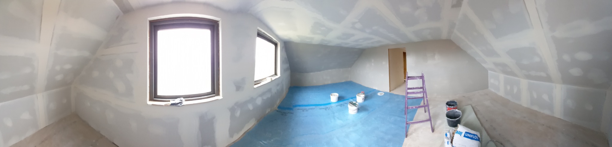 Poddasze w Dominowie w trakcie szpachlowania: widoczne okna, ściany i sufit pokryte gładzią szpachlową, podłoga zabezpieczona folią, drabina i wiaderka z materiałami.