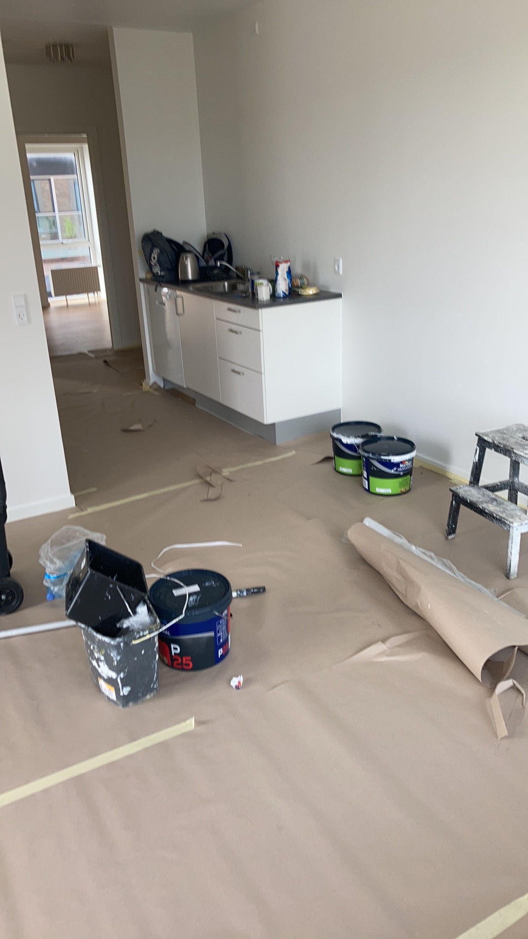Przygotowania do malowania mieszkania: zabezpieczona folią podłoga, wiadro z farbą, pędzle i drabina w pustym pomieszczeniu z aneksem kuchennym.