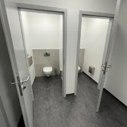 Dwa otwarte wejścia do czystych, minimalistycznych toalet w biurze, widok z korytarza na szarą podłogę i białe ściany.