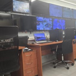 Omega Balcerak Sp. z o.o. - Stanowiska pracy z monitorami CCTV w pomieszczeniu kontroli. Ekrany pokazują obraz z kamer. Biurka z komputerami i krzesłami. Widok na salę monitoringu.