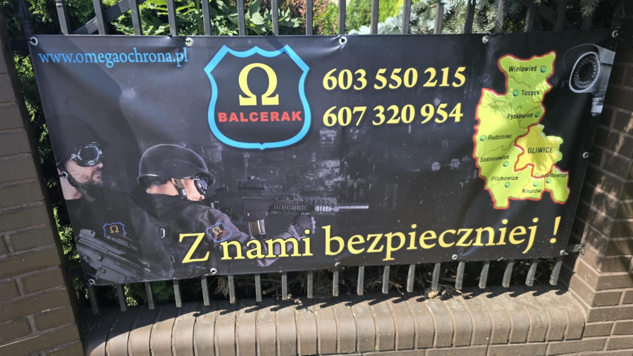 Baner reklamowy z hasłem 'Z nami bezpieczniej!', prezentujący ofertę. Na banerze umundurowani ochroniarze z bronią, logo firmy i kontakt. Widoczna mapa z lokalizacjami.