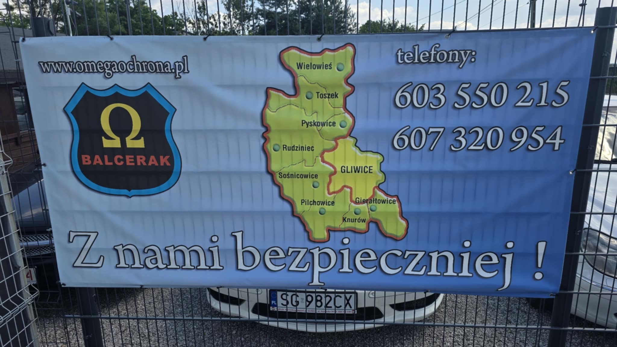 Baner z logo firmy ochroniarskiej Omega Ochrona i numerami telefonów, umieszczony na ogrodzeniu z widocznym fragmentem samochodu w tle.