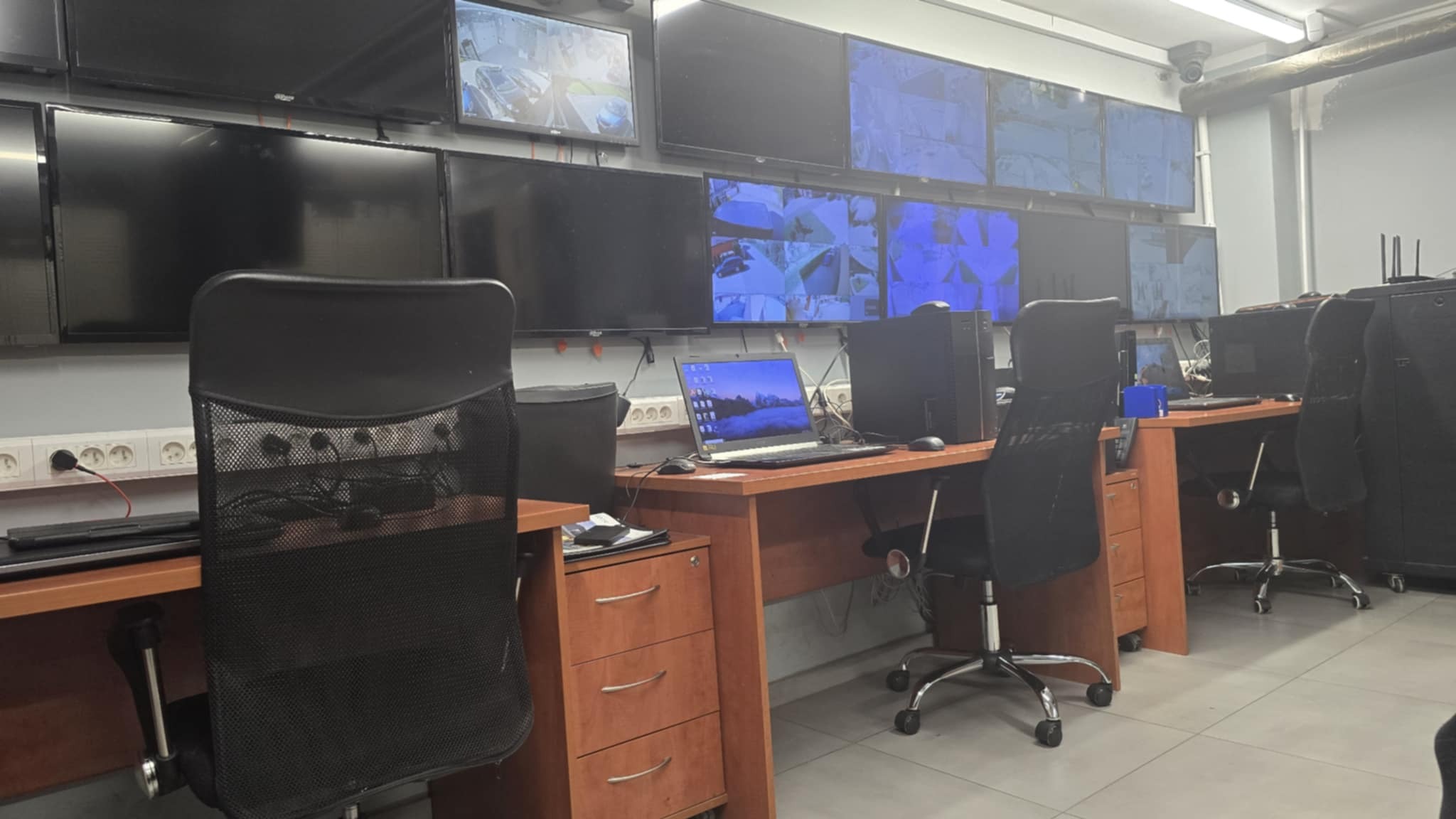 Stanowiska pracy z monitorami CCTV w pomieszczeniu kontroli. Ekrany pokazują obraz z kamer. Biurka z komputerami i krzesłami. Widok na salę monitoringu.