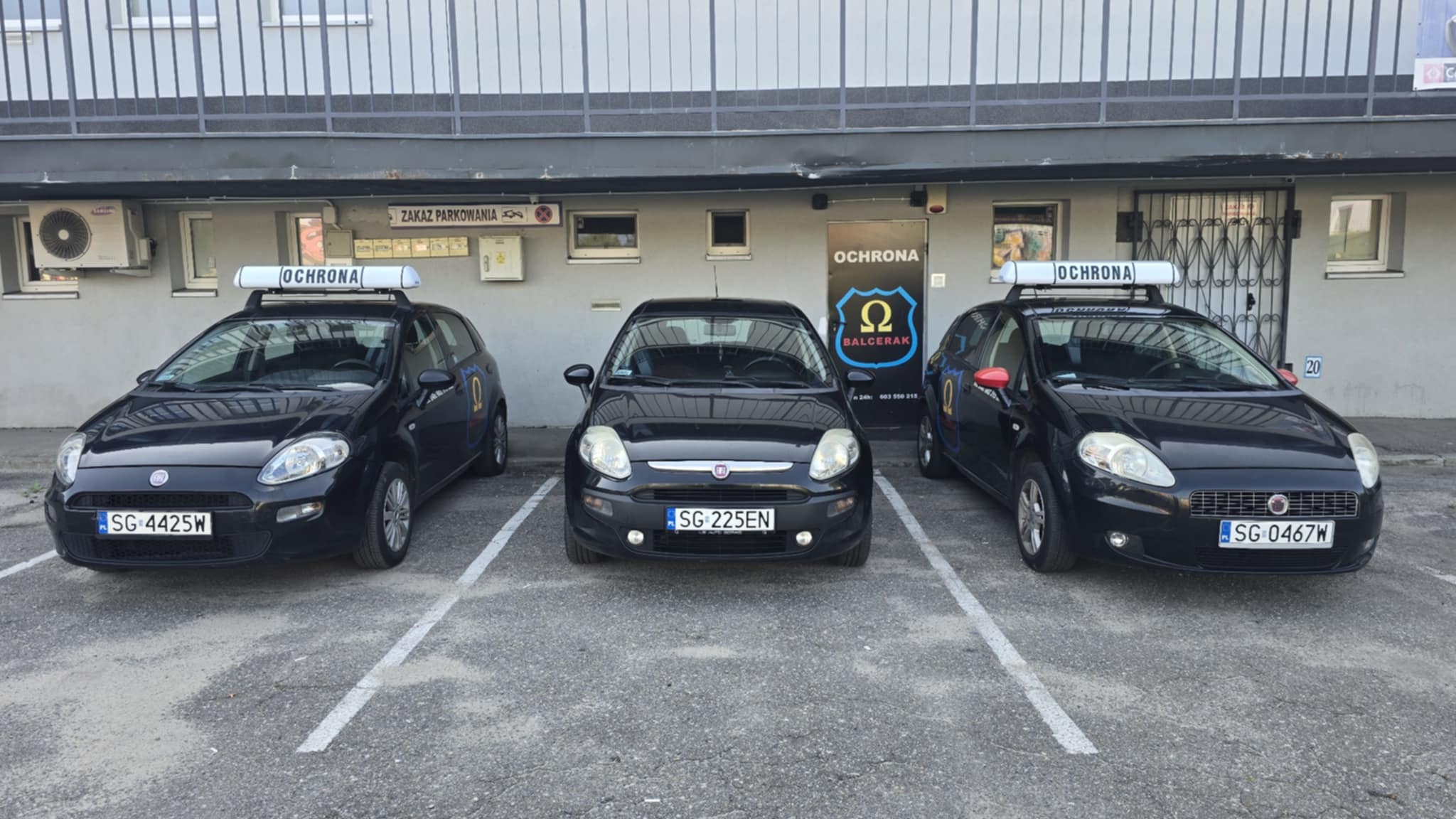 Trzy czarne samochody z napisem 'Ochrona' na dachu zaparkowane przed budynkiem z logo firmy ochroniarskiej 'Balcerak'.