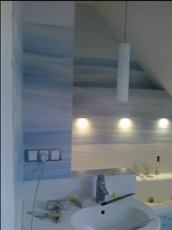 Błękitna marmurkowa okładzina ściany łazienki z zamontowanymi gniazdkami elektrycznymi, widoczna umywalka z baterią, narzędzia glazurnicze na blacie i cylindryczna lampa wisząca.