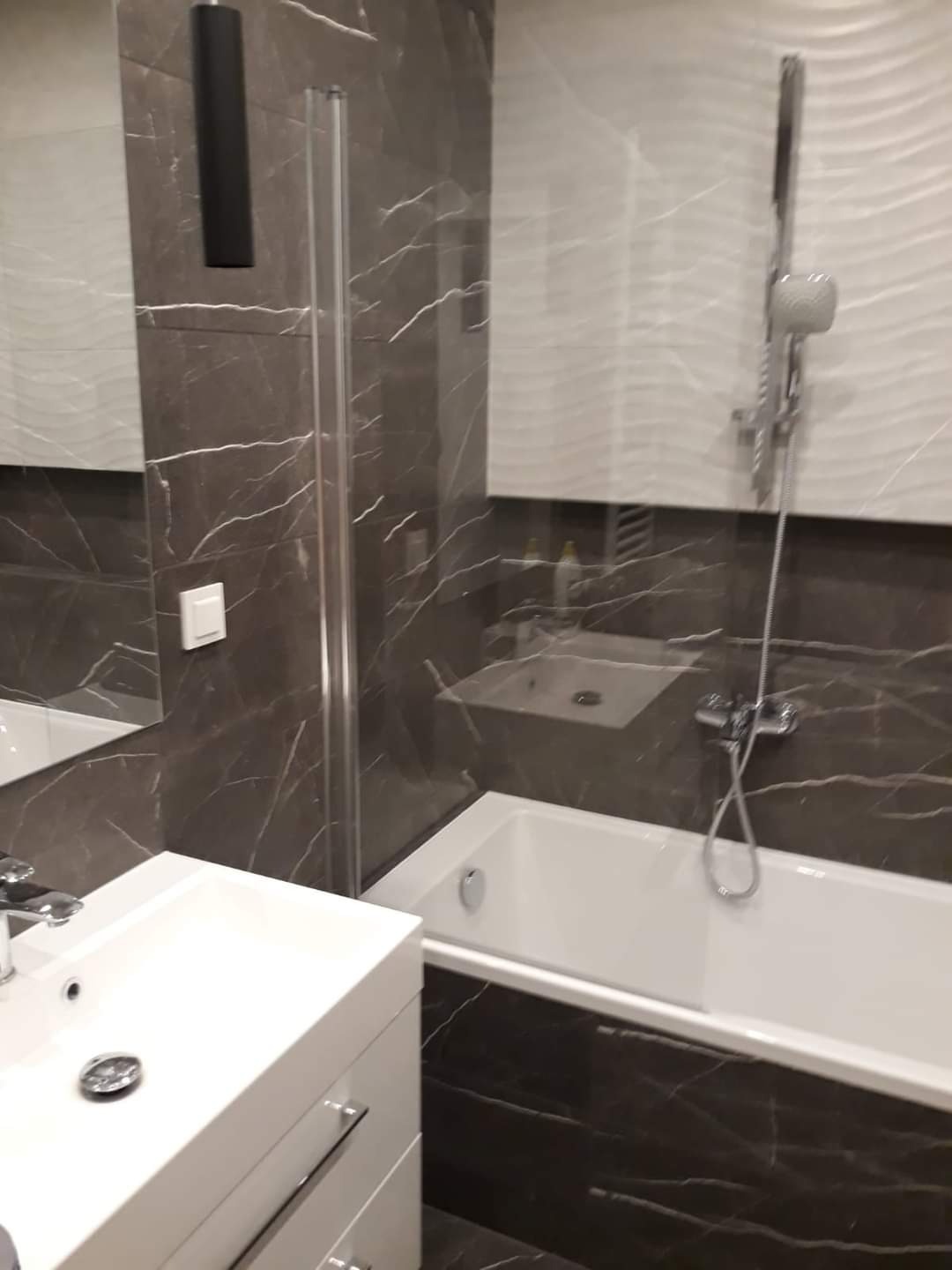 Wykończona łazienka z marmurowymi płytkami, białą wanną i umywalką z szafką, widoczne detale armatury i oświetlenia.