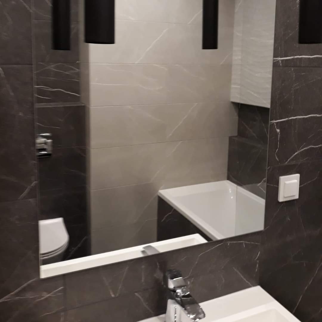 Łazienka wykończona w szarościach i czerni z marmurkowym wzorem na płytkach, widoczna umywalka z chromowaną baterią oraz odbicie toalety i wanny w lustrze.
