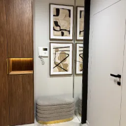 Nowoczesne wykończenie przedpokoju w Krakowie: zabudowa szafy z oświetloną niszą, tapicerowany puf z elementami złota, abstrakcyjne obrazy na ścianie i duże lustro.