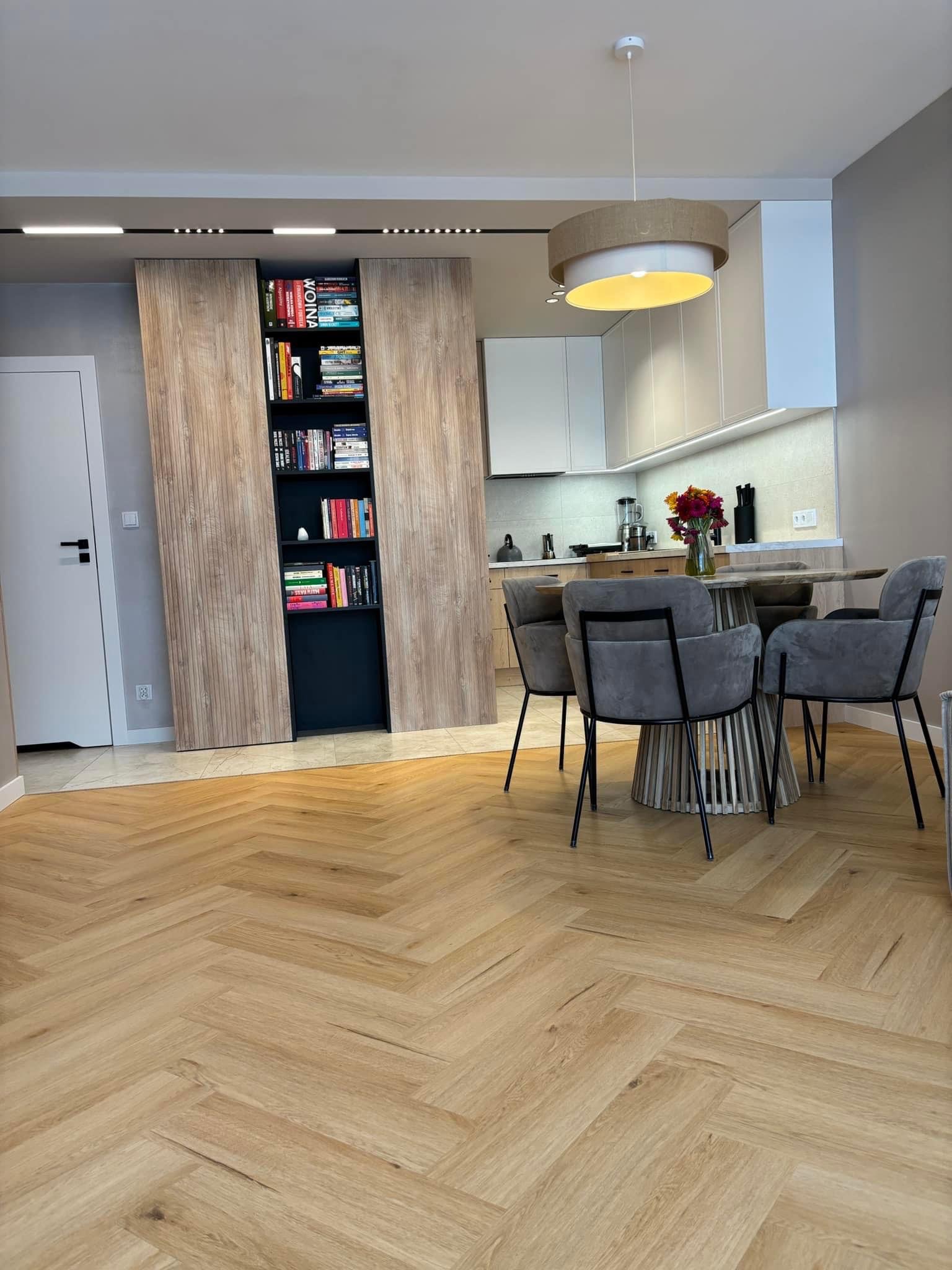Jasny parkiet ułożony w jodełkę francuską, widok na stół z szarymi krzesłami, zabudowę kuchenną i regał na książki z drewnianymi przesuwnymi drzwiami.