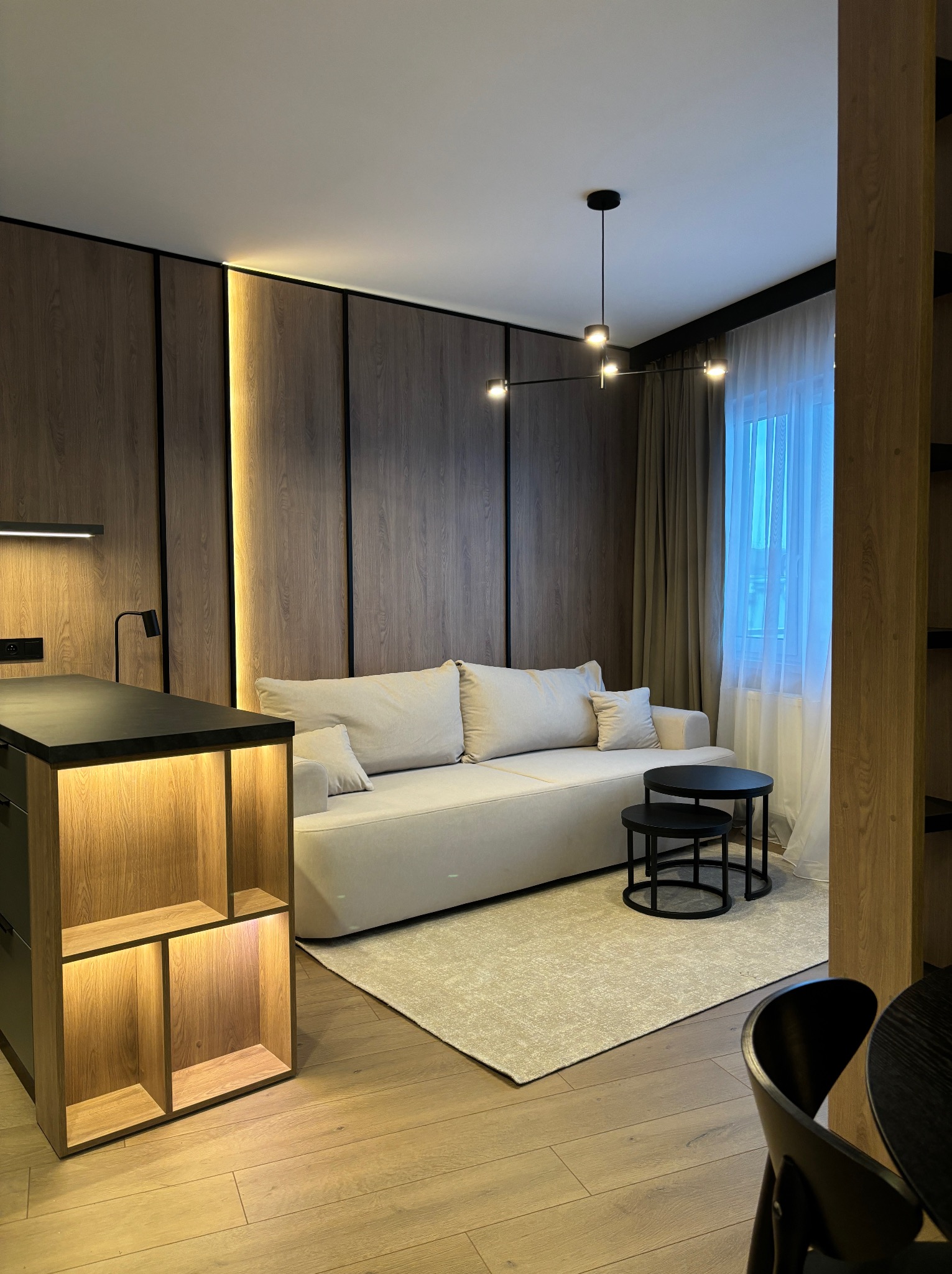 Jasny salon z drewnianymi panelami ściennymi z podświetleniem LED, beżową sofą, czarnymi stolikami kawowymi i nowoczesną lampą sufitową.