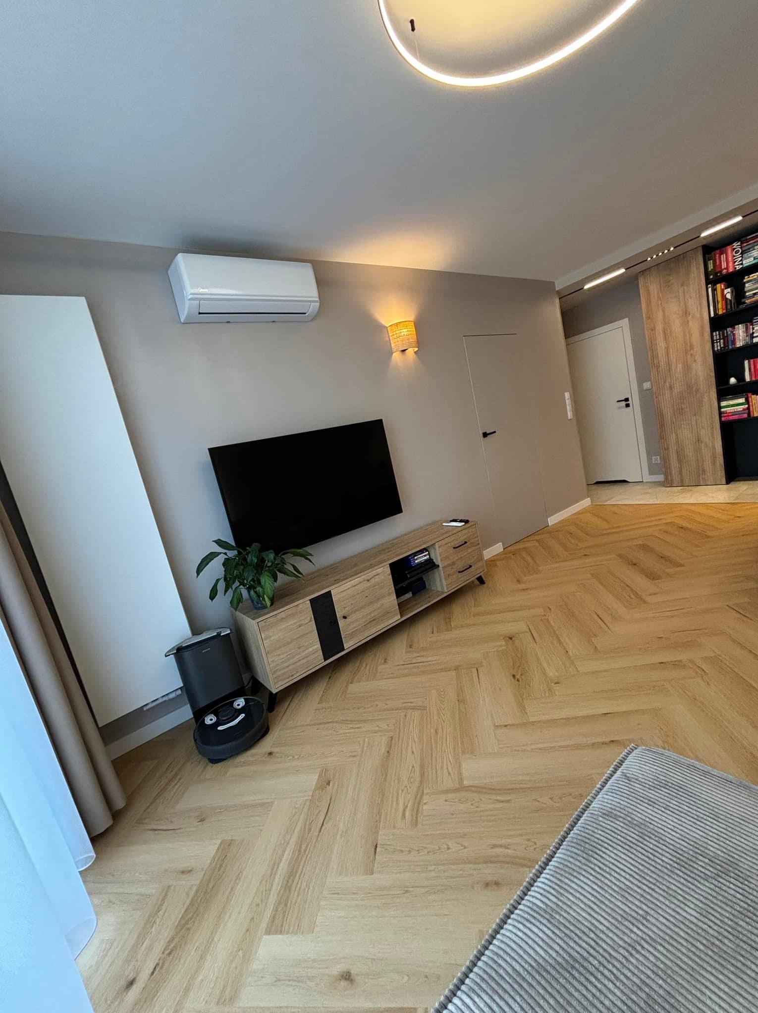 Nowoczesny salon z parkietem w jodełkę, szarą ścianą i minimalistycznymi meblami, robot sprzątający parkuje obok rośliny doniczkowej.