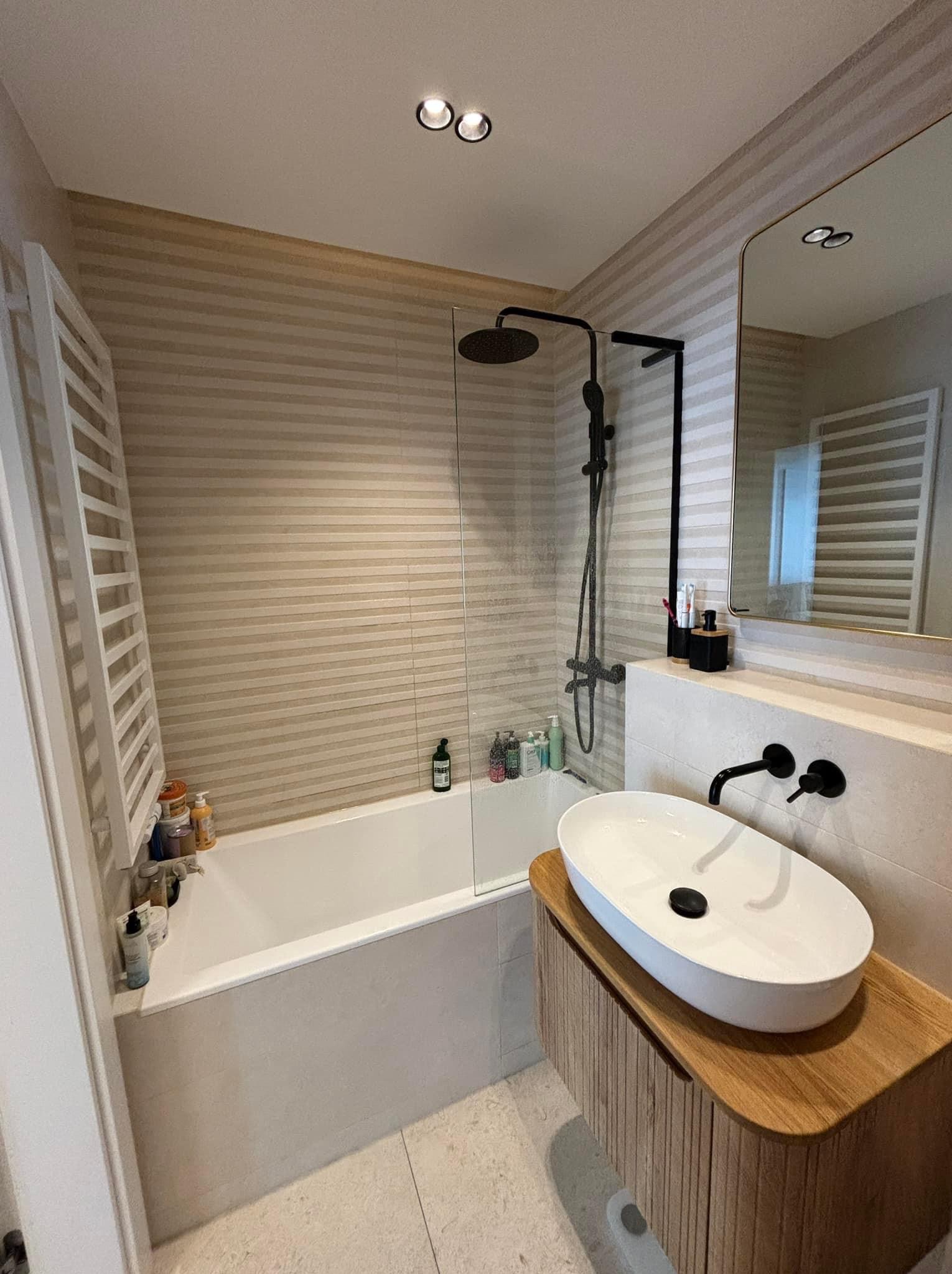 Nowoczesna łazienka z wanną za szklanym panelem, czarną armaturą i umywalką nablatową na drewnianej szafce z ryflowanym frontem, ściany wykończone płytkami w poziome pasy.